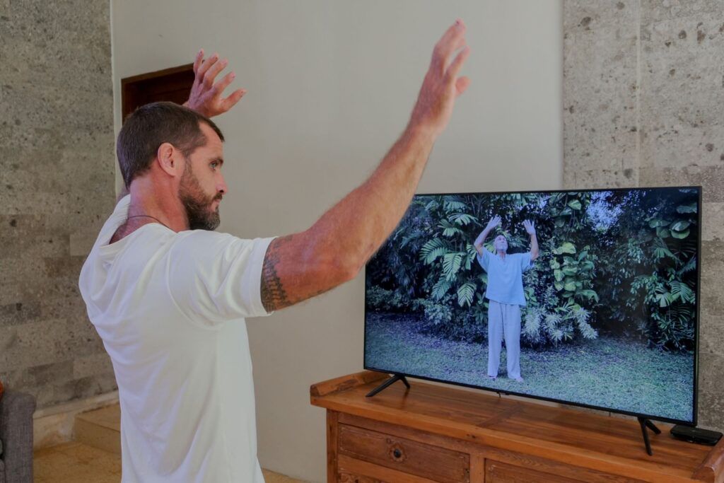 A man practicing Qigong indoors while following an online video lesson displayed on a television screen.