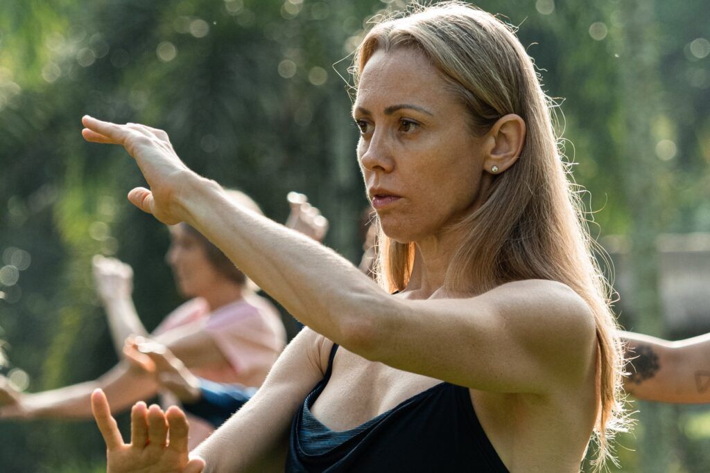 A woman practicing Qigong outdoors, focusing on slow, mindful arm movements during a group session.