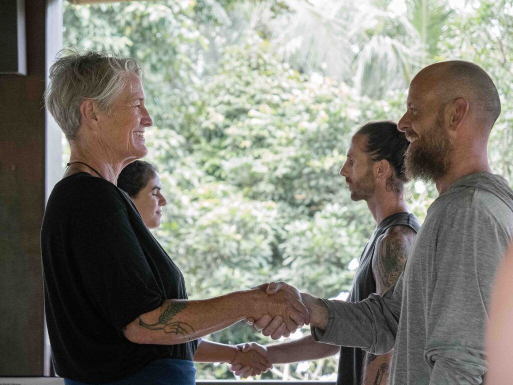 Two Qigong students warmly shaking hands after a practice session in a natural outdoor setting.
