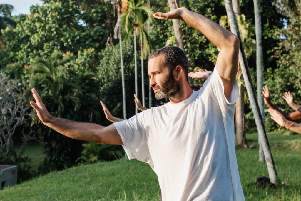 Man practicing Qigong outdoors with extended arms, performing a flowing movement in a natural garden setting.